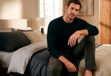 Como Ser um Homem Bom de Cama: O Guia Definitivo para Surpreender Homem confiante sentado na beira da cama em um quarto com iluminação aconchegante, representando o conceito de bom de cama.